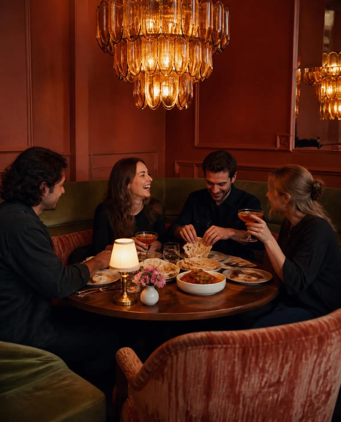 Vier Gäste sitzen abends an einem runden Tisch, lachen, teilen Speisen und halten Cocktails in der Hand